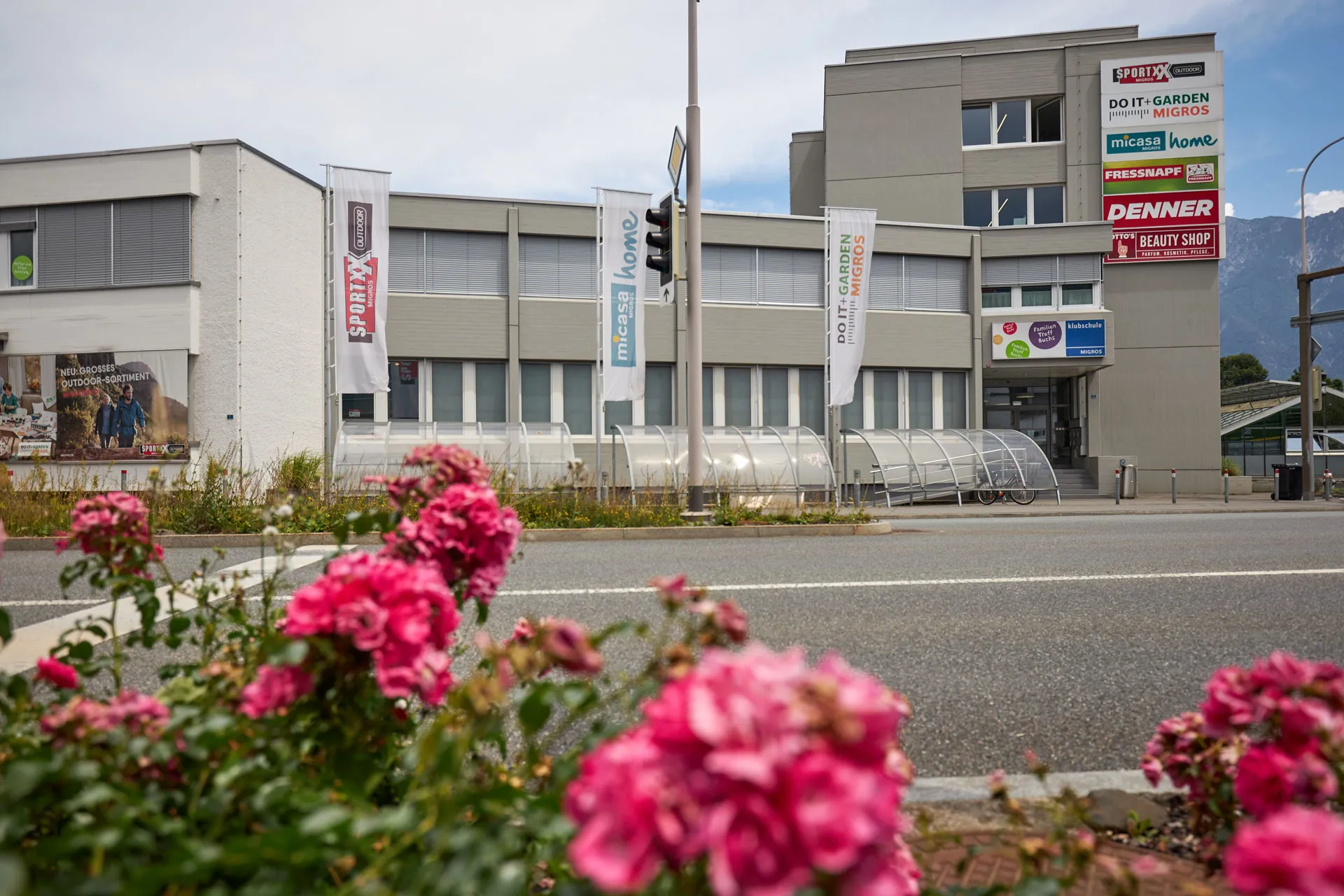 Aussenansicht Gebäudes Klubschule Buchs, umgeben von Strasse und pinkfarbenen Blumen