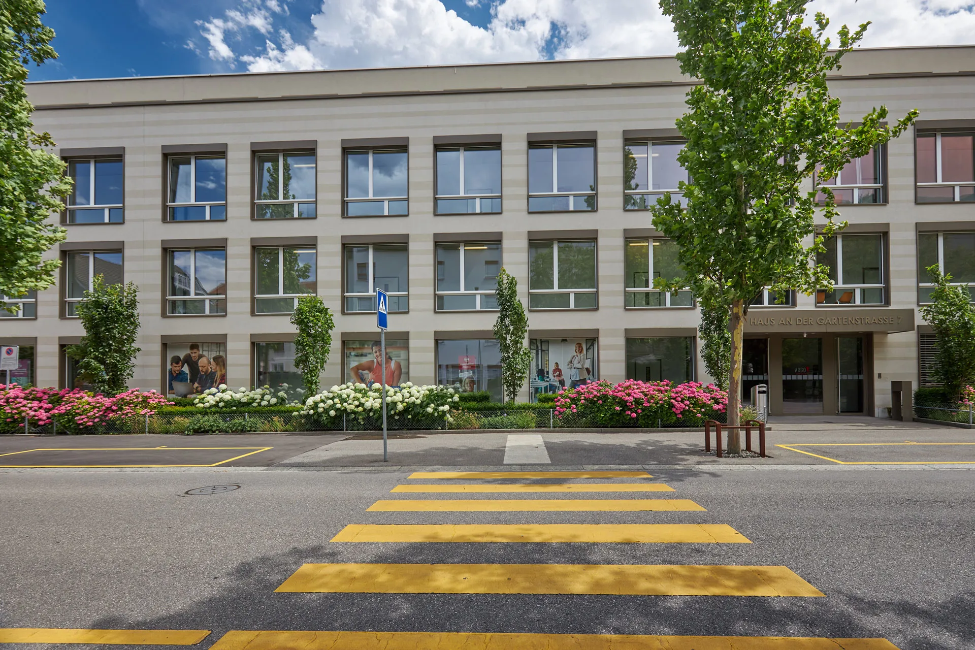 Aussenansicht Gebäude Klubschule Chur mit Fussgängerstreifen, Bäumen und Blumen